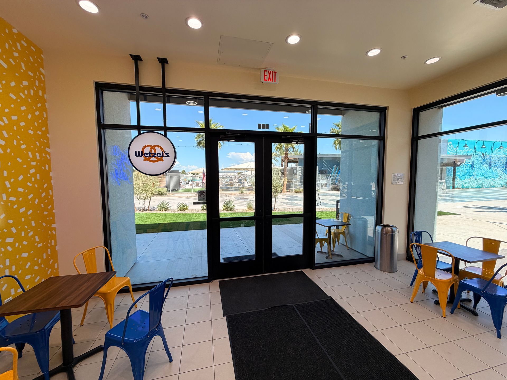 Empty café entrance with tables, chairs, and large glass doors, sunlight streaming in.