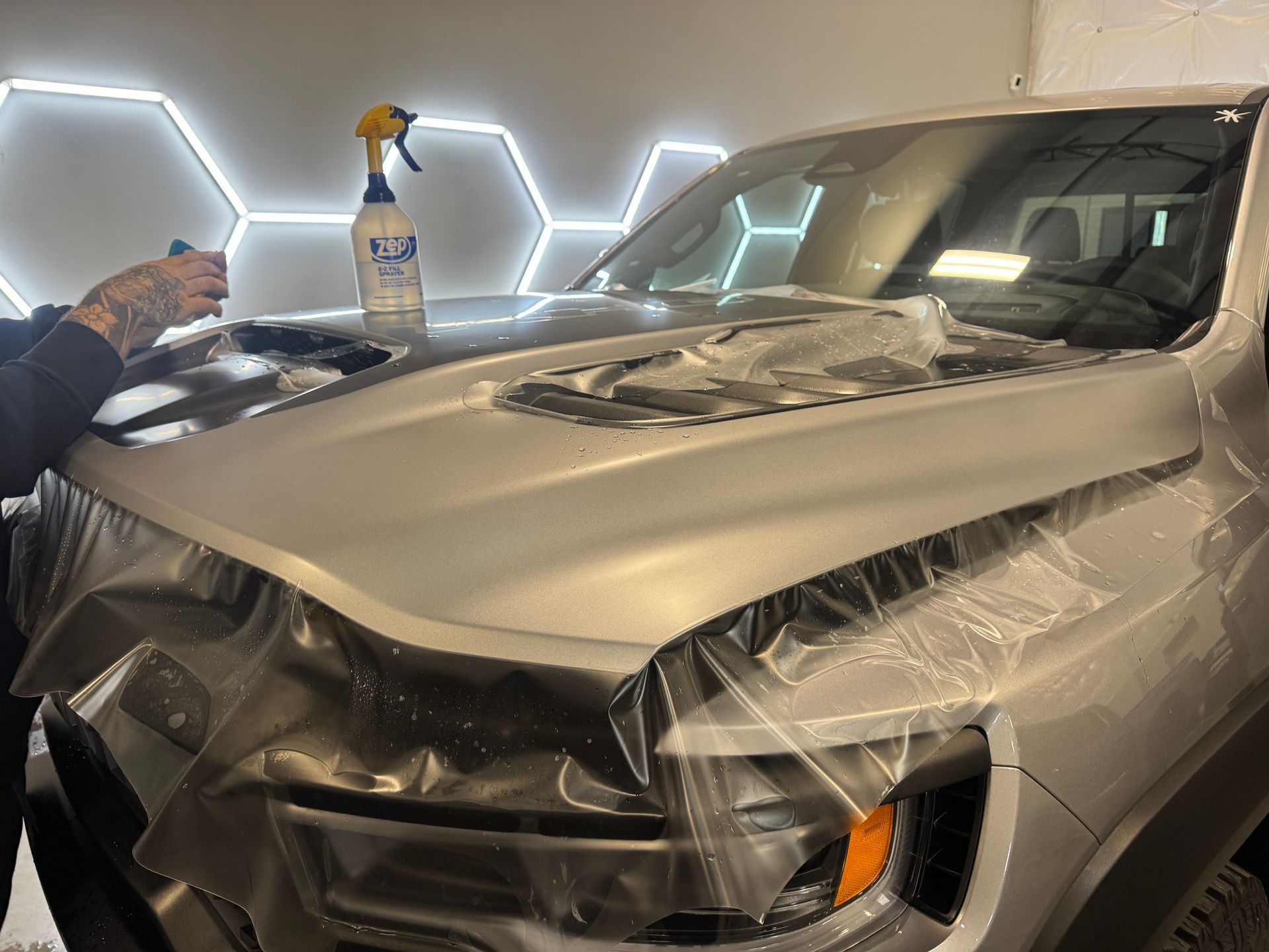 Silver car wrapped in clear plastic in a garage, with a spray bottle and a person’s hand on the left.