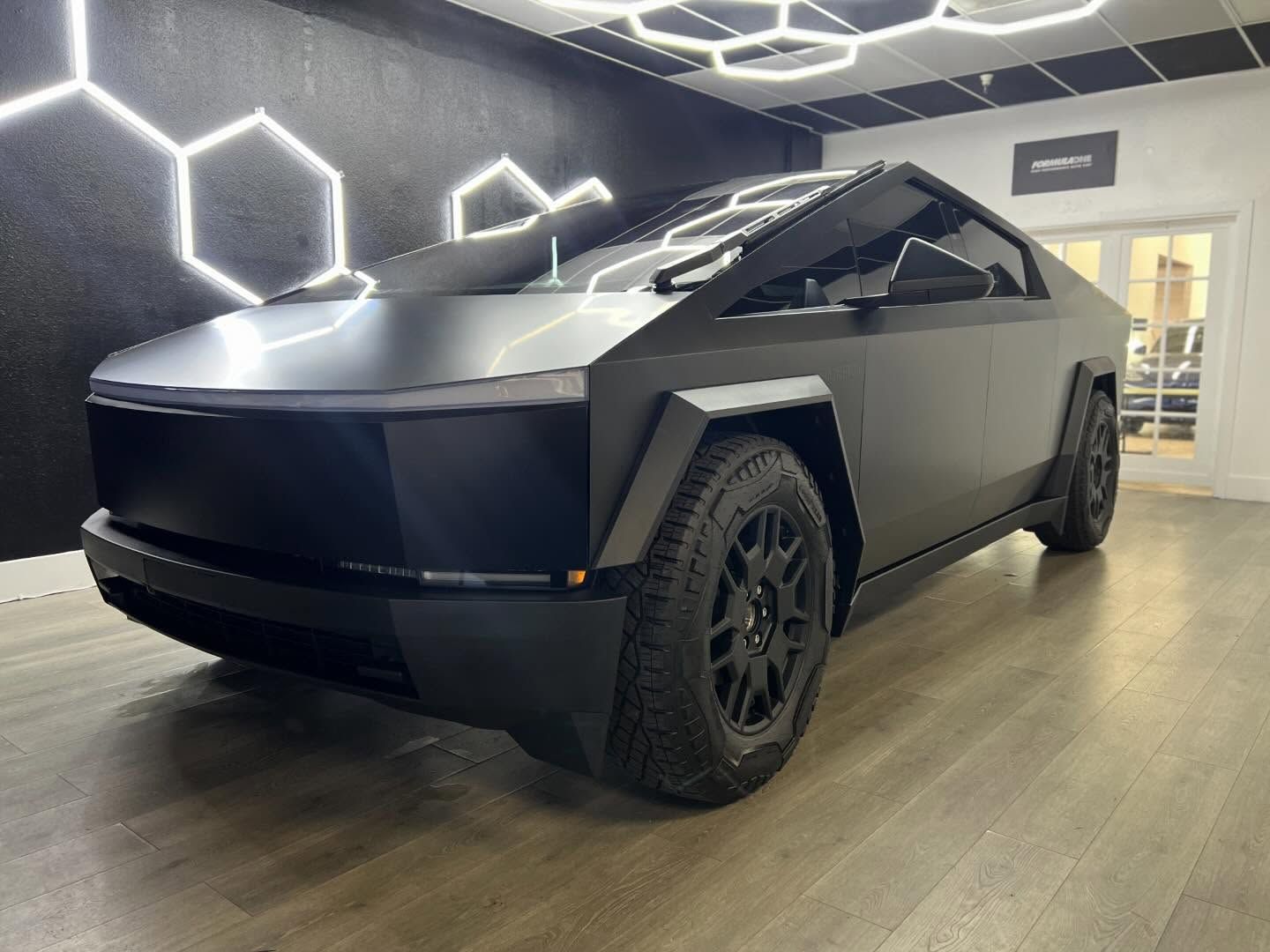 Matte black Cybertruck displayed indoors on a showroom floor with geometric wall lights
