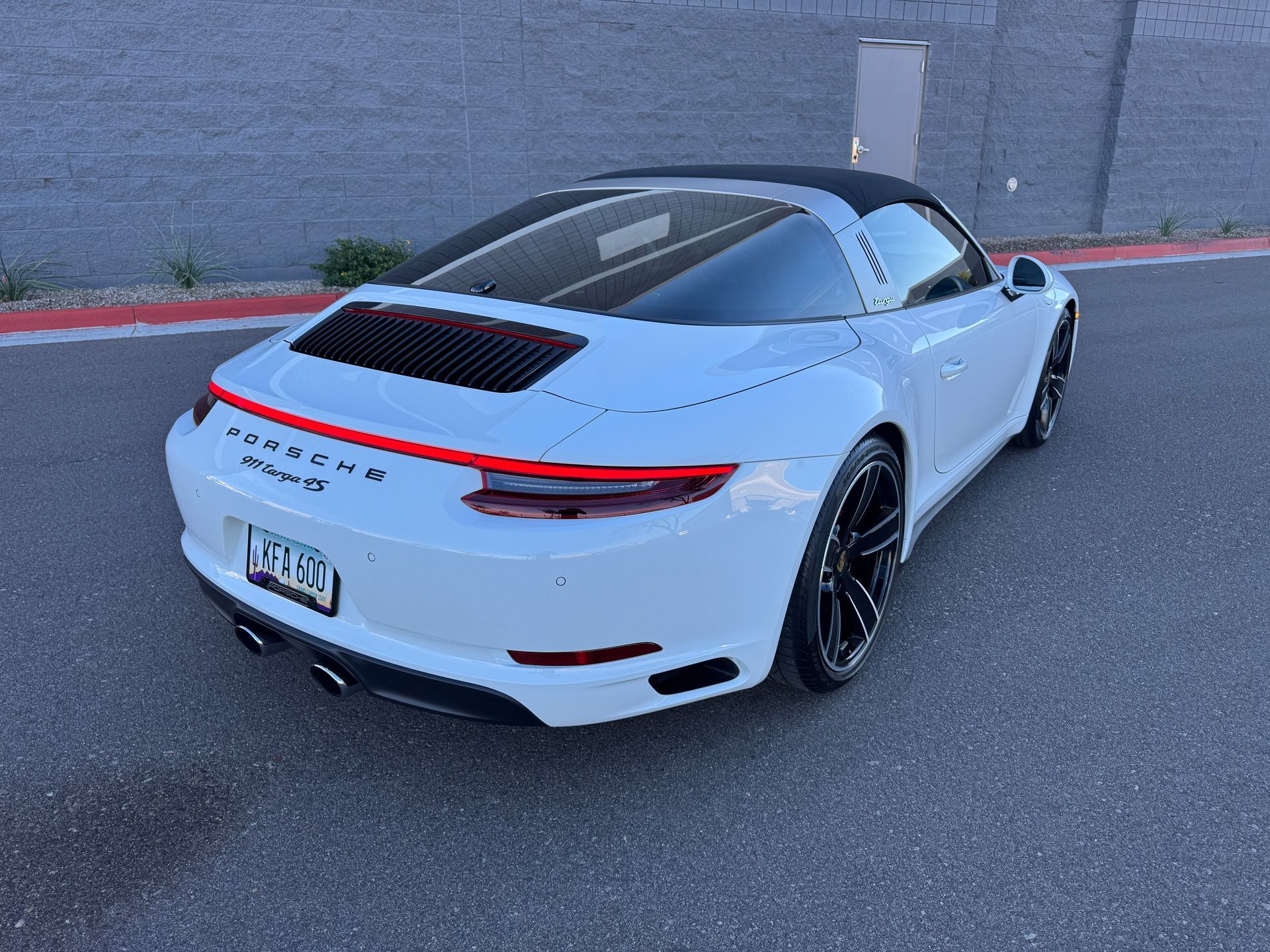 White Porsche 911 parked on a street, rear three-quarter view with a gray wall background