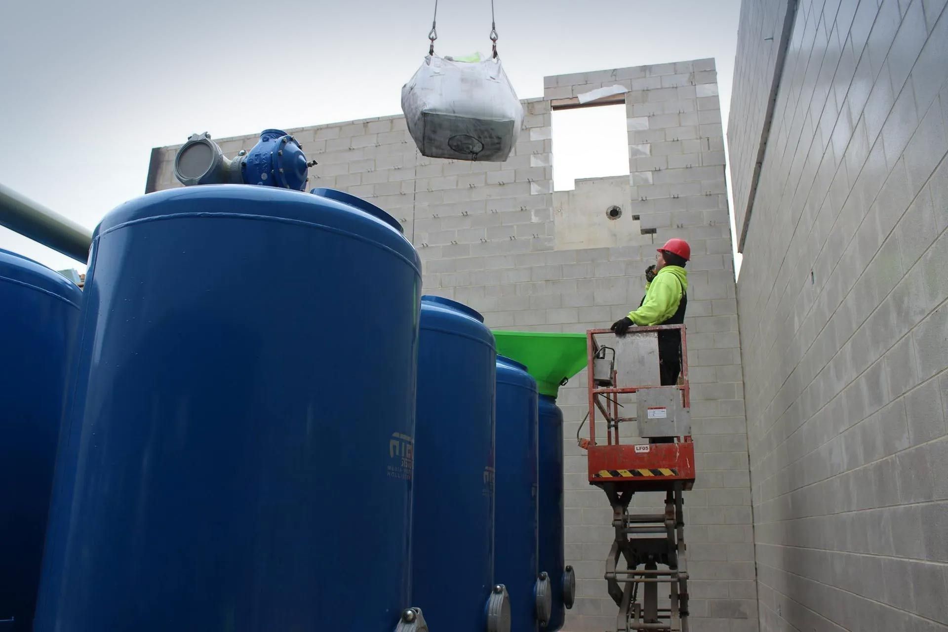 A man is standing on a lift in front of a row of blue tanks.