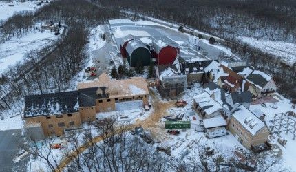 Aerial view of a snowy construction site. Buildings with varying roof colors and construction materials are surrounded by snow. 