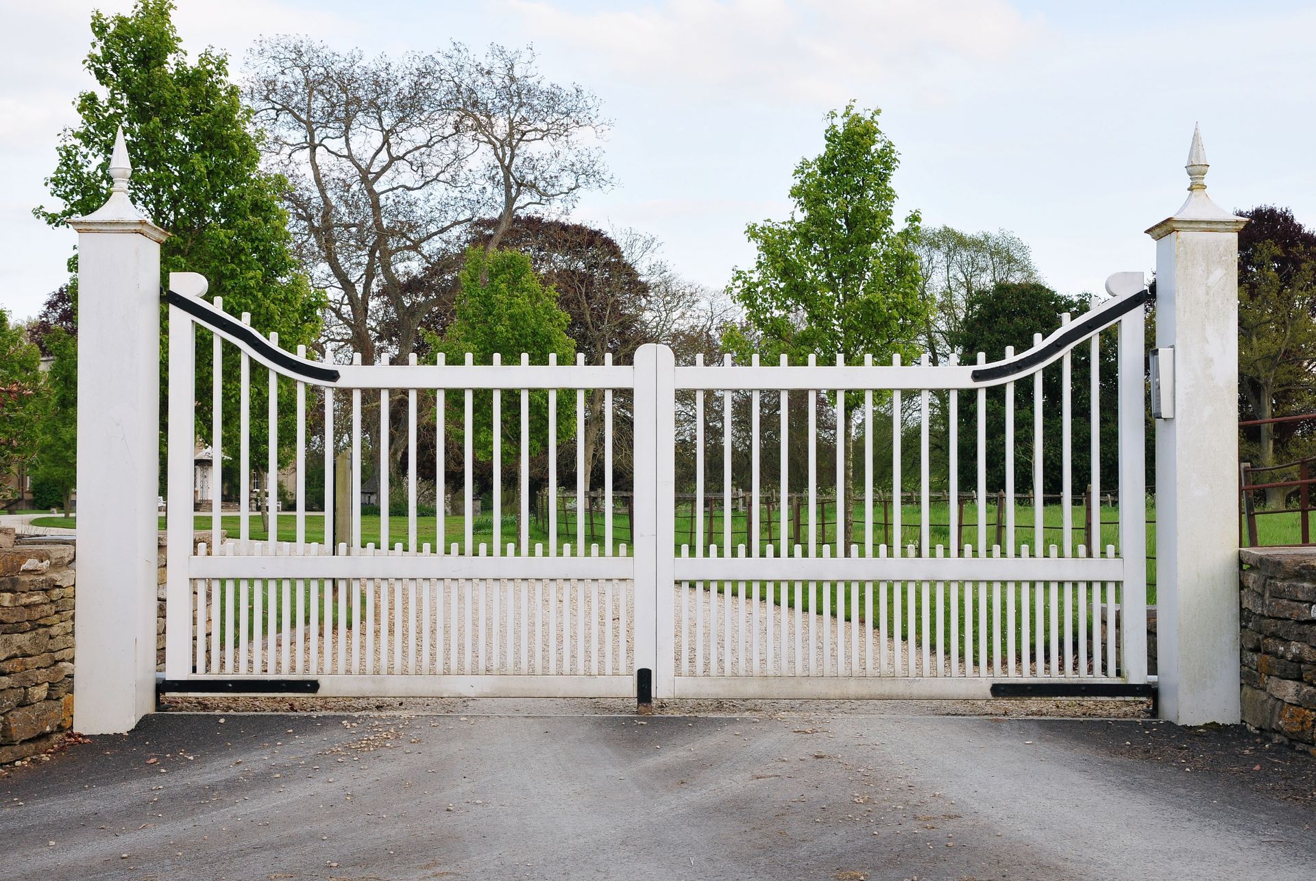 White metal driveway gate with arched top between stone pillars, set before trees and a road.