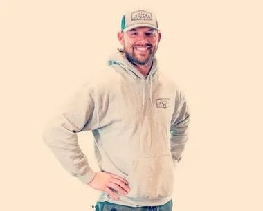 Smiling man in a light hoodie and cap, standing with hands on hips against a plain background