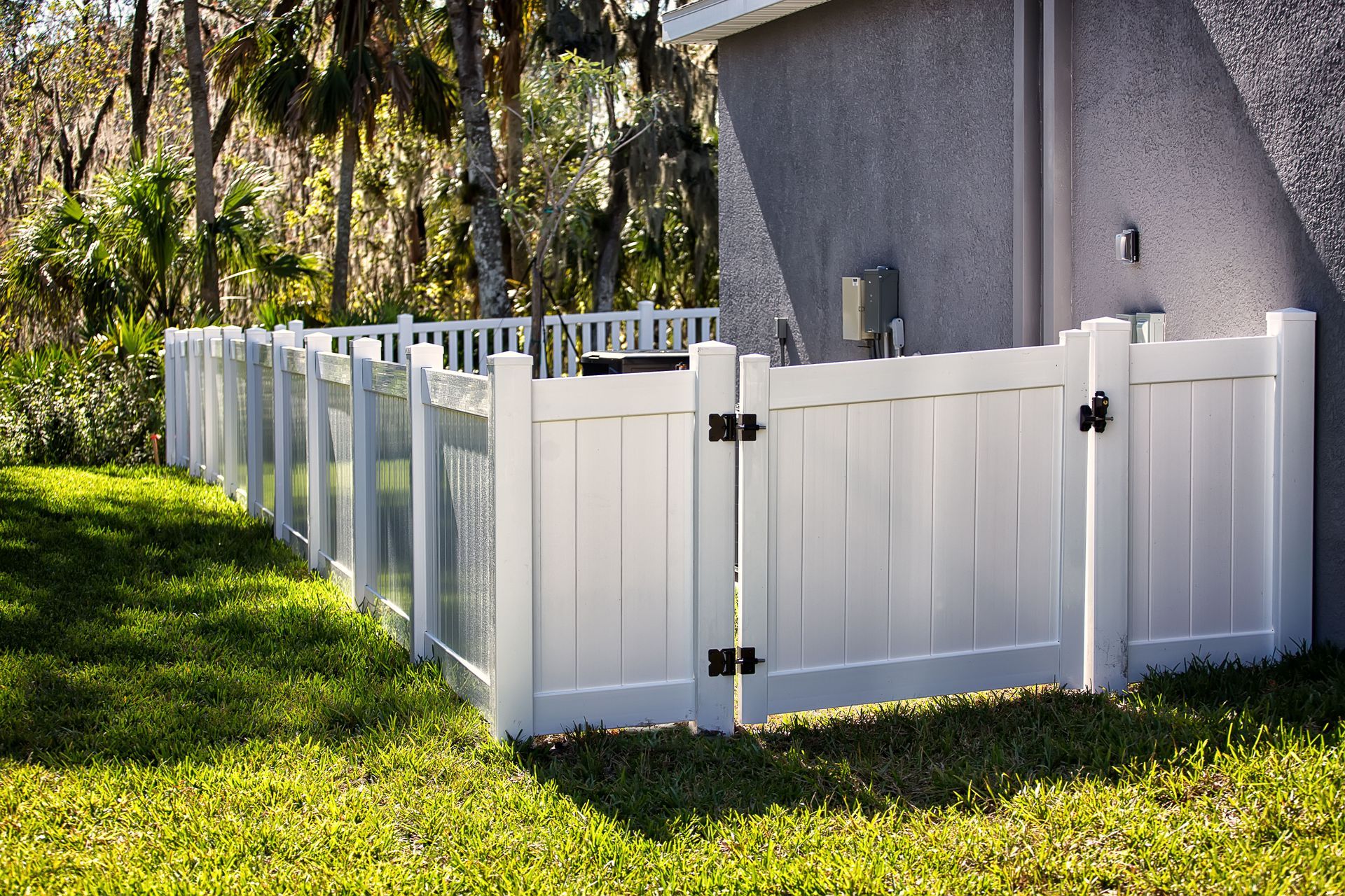 White vinyl fence along a grassy yard beside a gray house and trees.