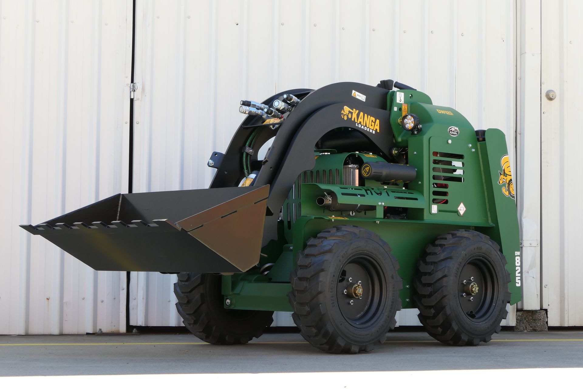 A green Kanga skid steer with a black bucket is parked in front of a white building