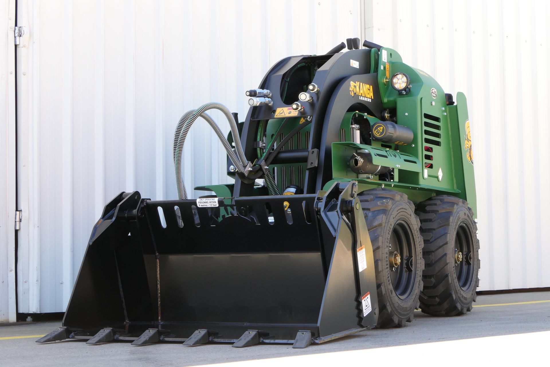 A green tractor with a black bucket is parked in front of a white wall.