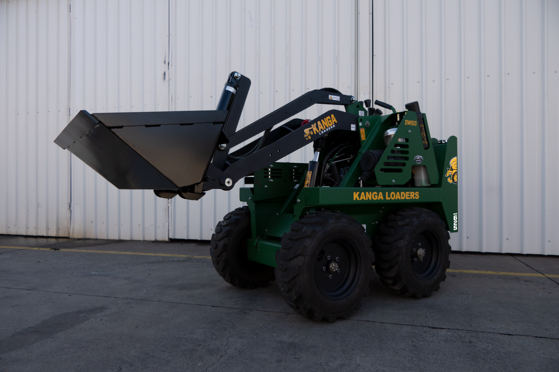 A green Kanga tractor with a black bucket is parked in front of a white building.