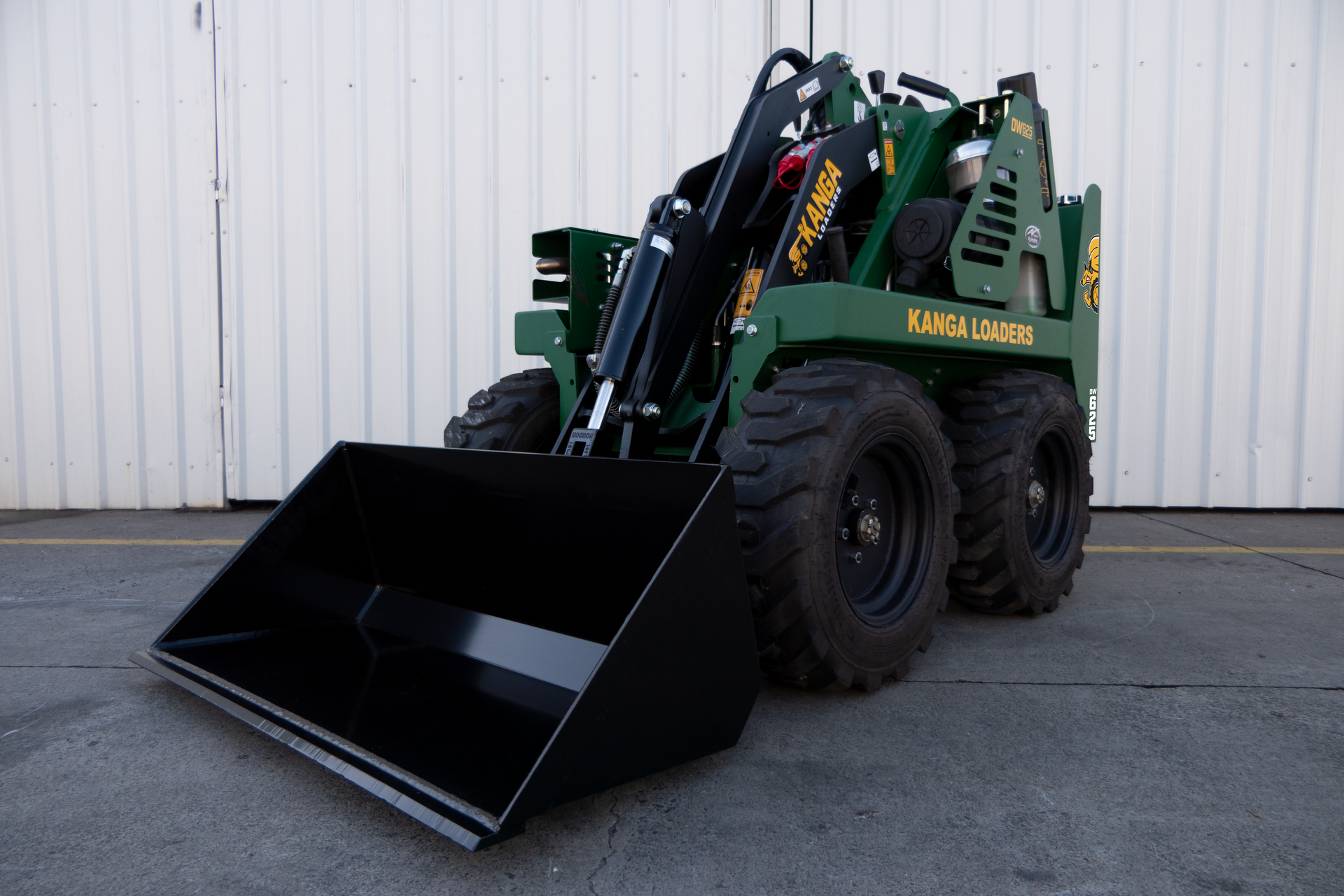 A green Kanga tractor with a black bucket is parked in front of a white wall.
