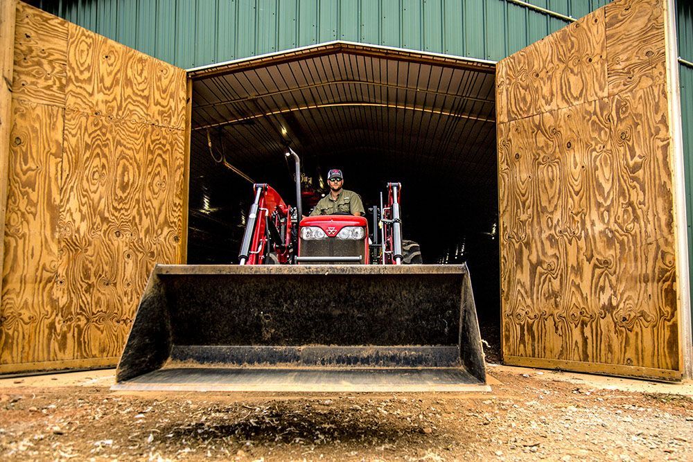 A man is driving a Massey Ferguson 2600H Series tractor in a garage with the doors open.