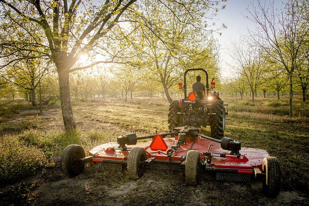 A man is driving a Massey Ferguson 2600H Series tractor through a field of trees.