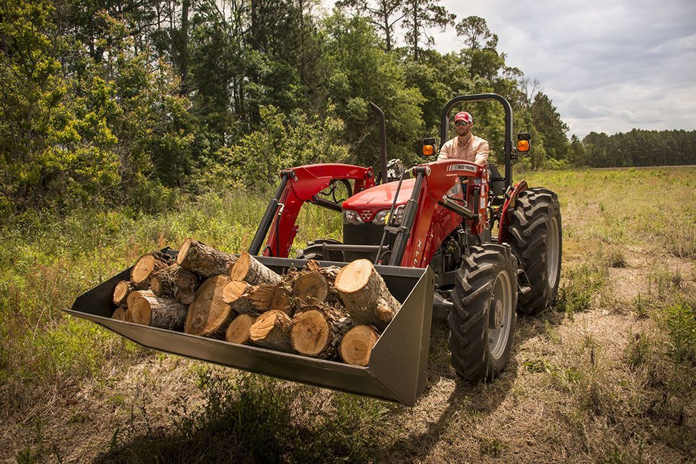 A man is driving a Massey Ferguson 2600H Series tractor with a bucket full of logs.