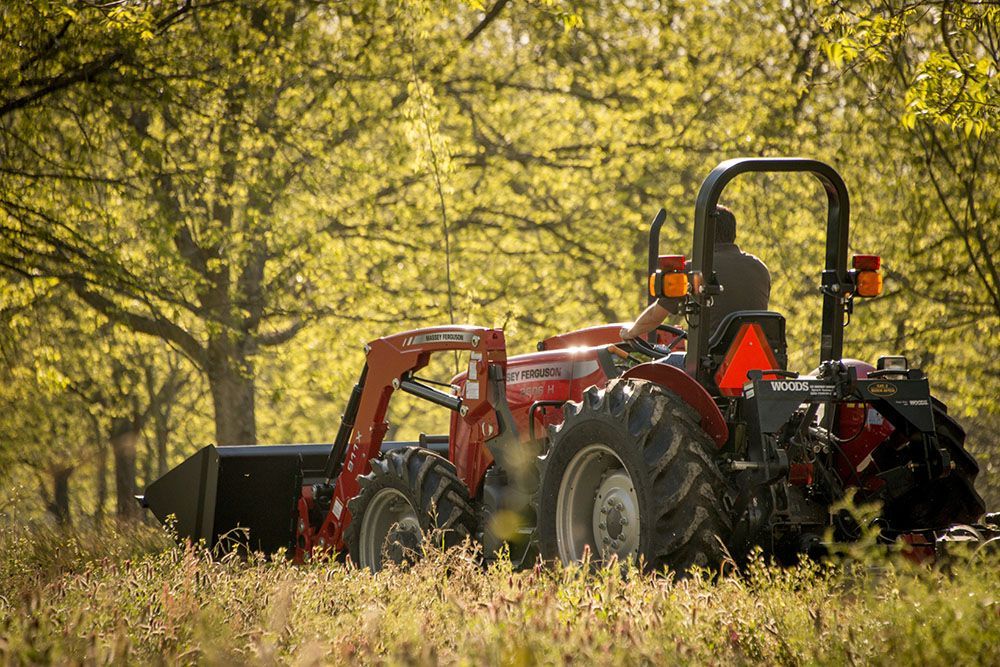 A red Massey Ferguson 2600H Series tractor is driving through a field with trees in the background.