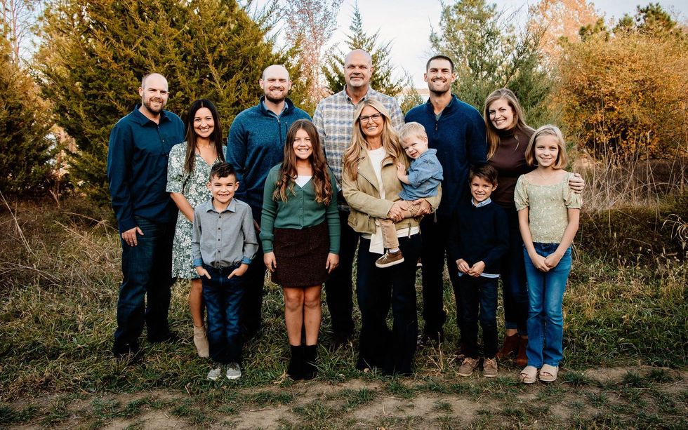 A large group poses for a portrait in a wooded, autumn outdoor setting. Everyone is smiling.