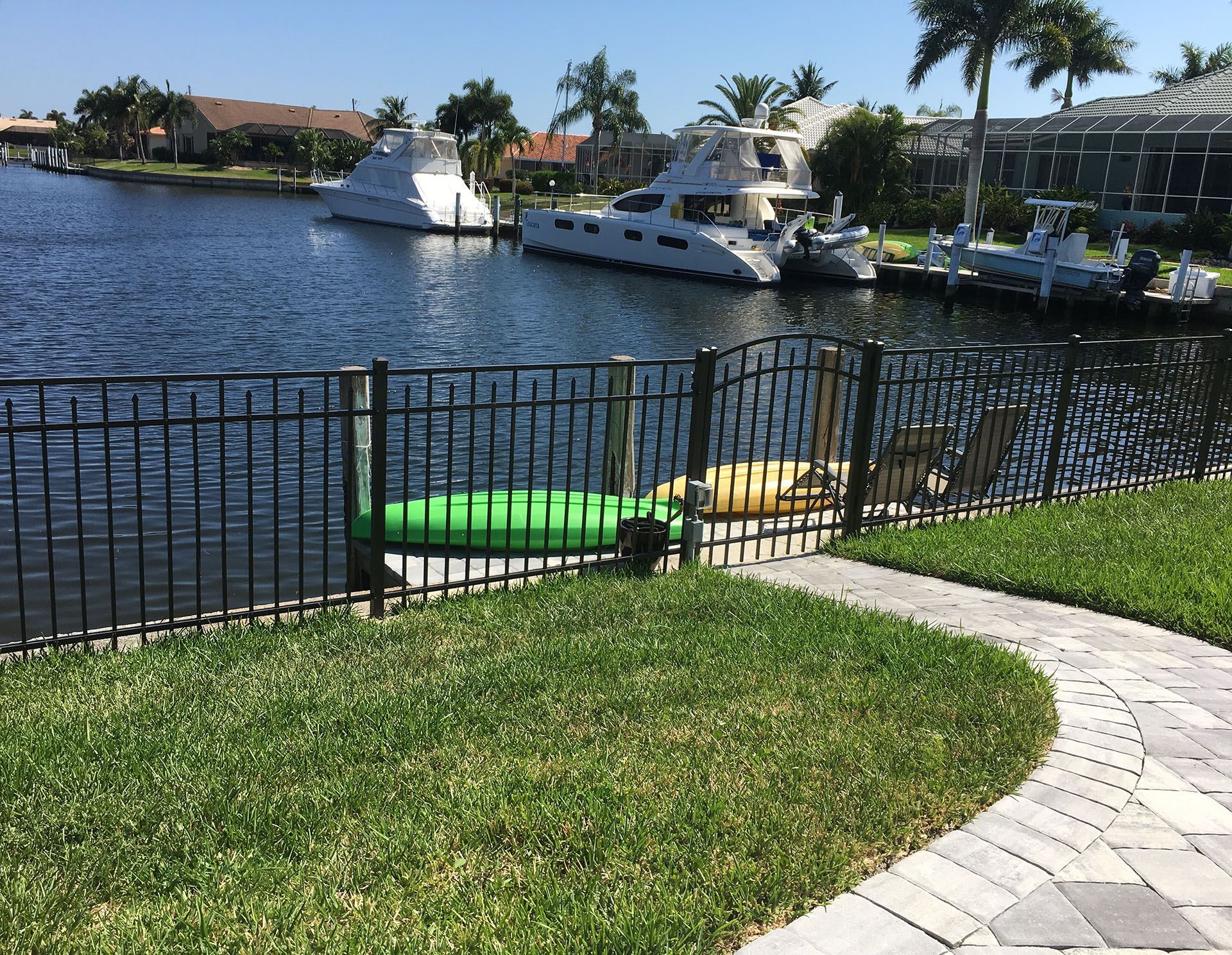 Black metal fence borders a grassy yard with a waterfront view. Boats are docked in the canal.