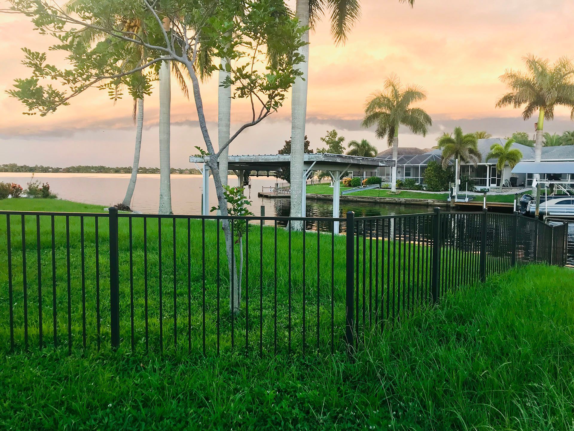 Black fence bordering lush green grass near a waterway with palm trees and houses during a sunset.