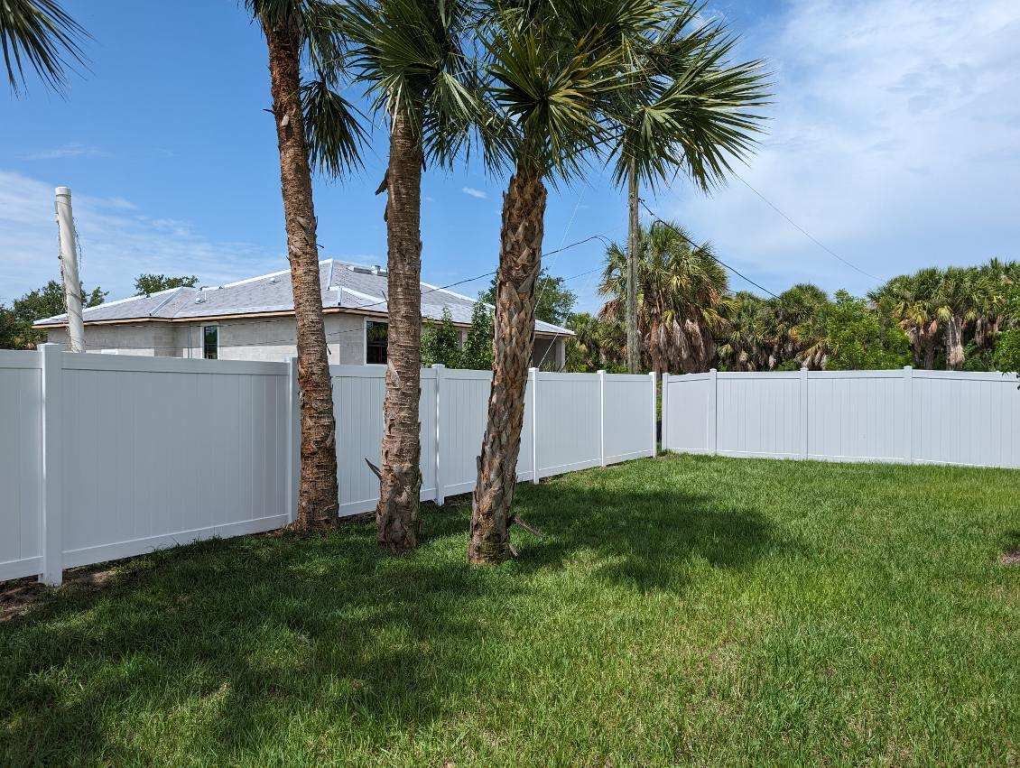 White fence surrounds a grassy yard with palm trees under a bright blue sky. A house is in the background.