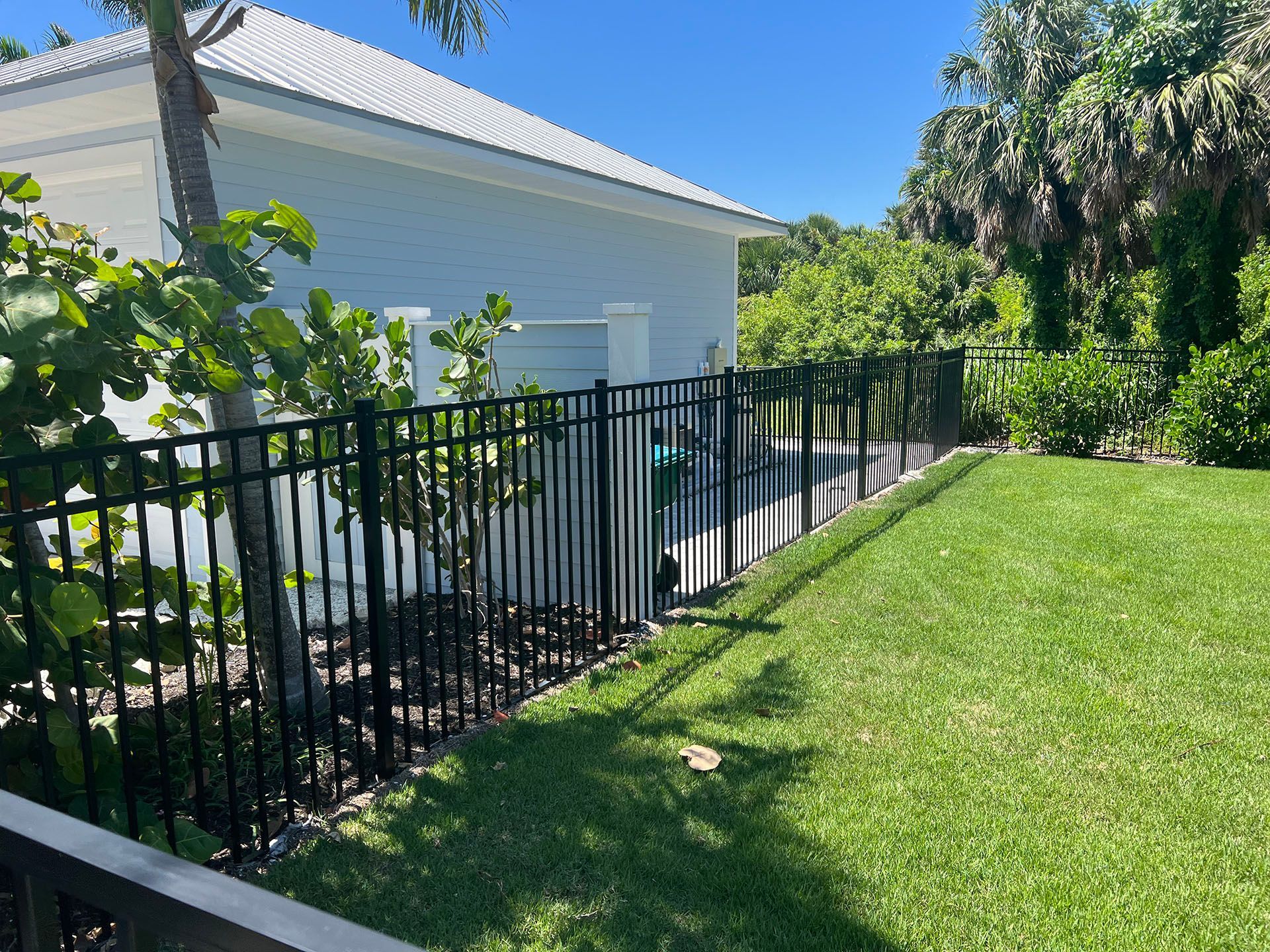 Black metal fence bordering a green lawn next to a light blue building and lush greenery. Sunny day.
