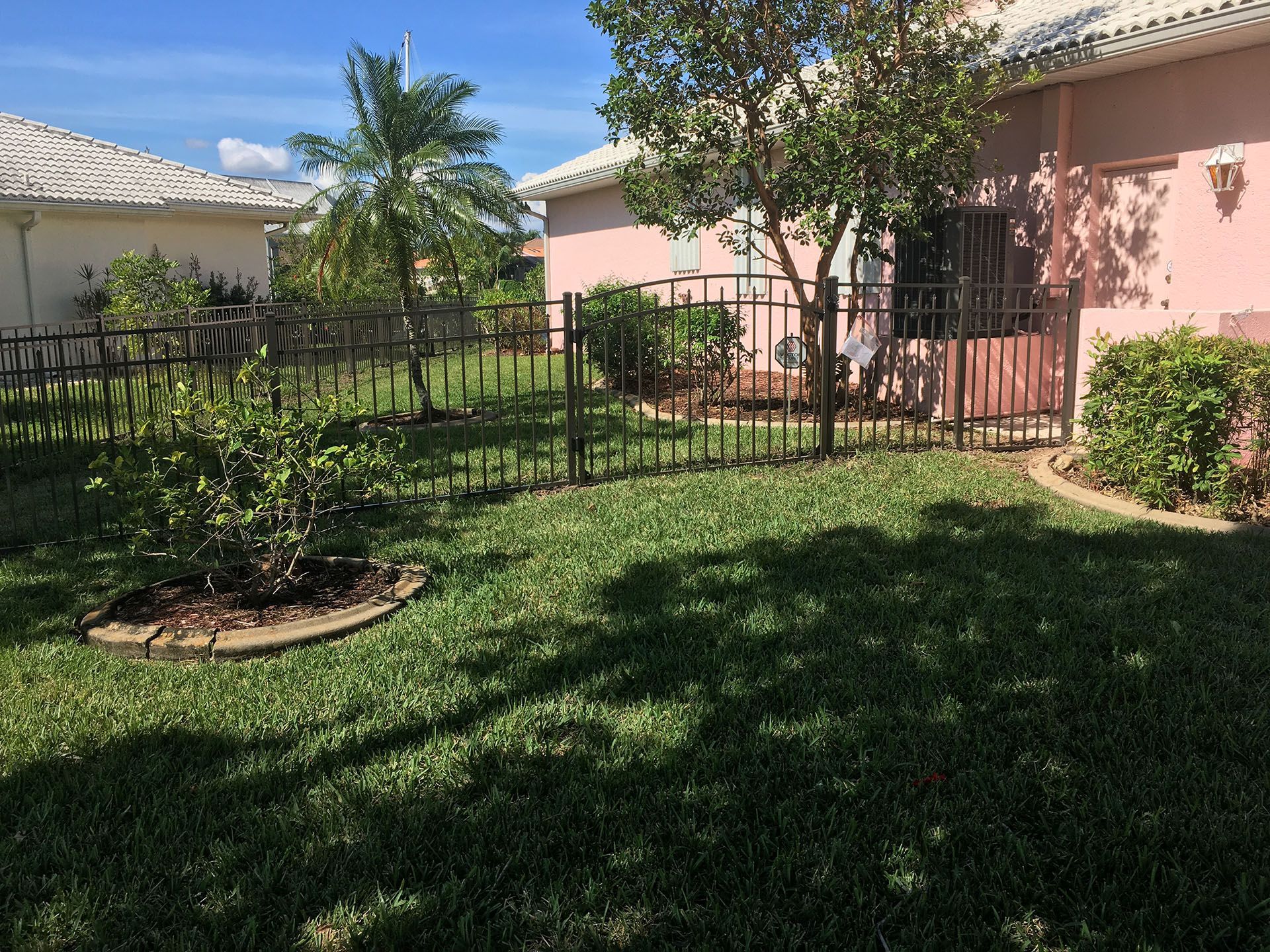 A fenced yard with green grass, small trees, and a pink house on a sunny day.