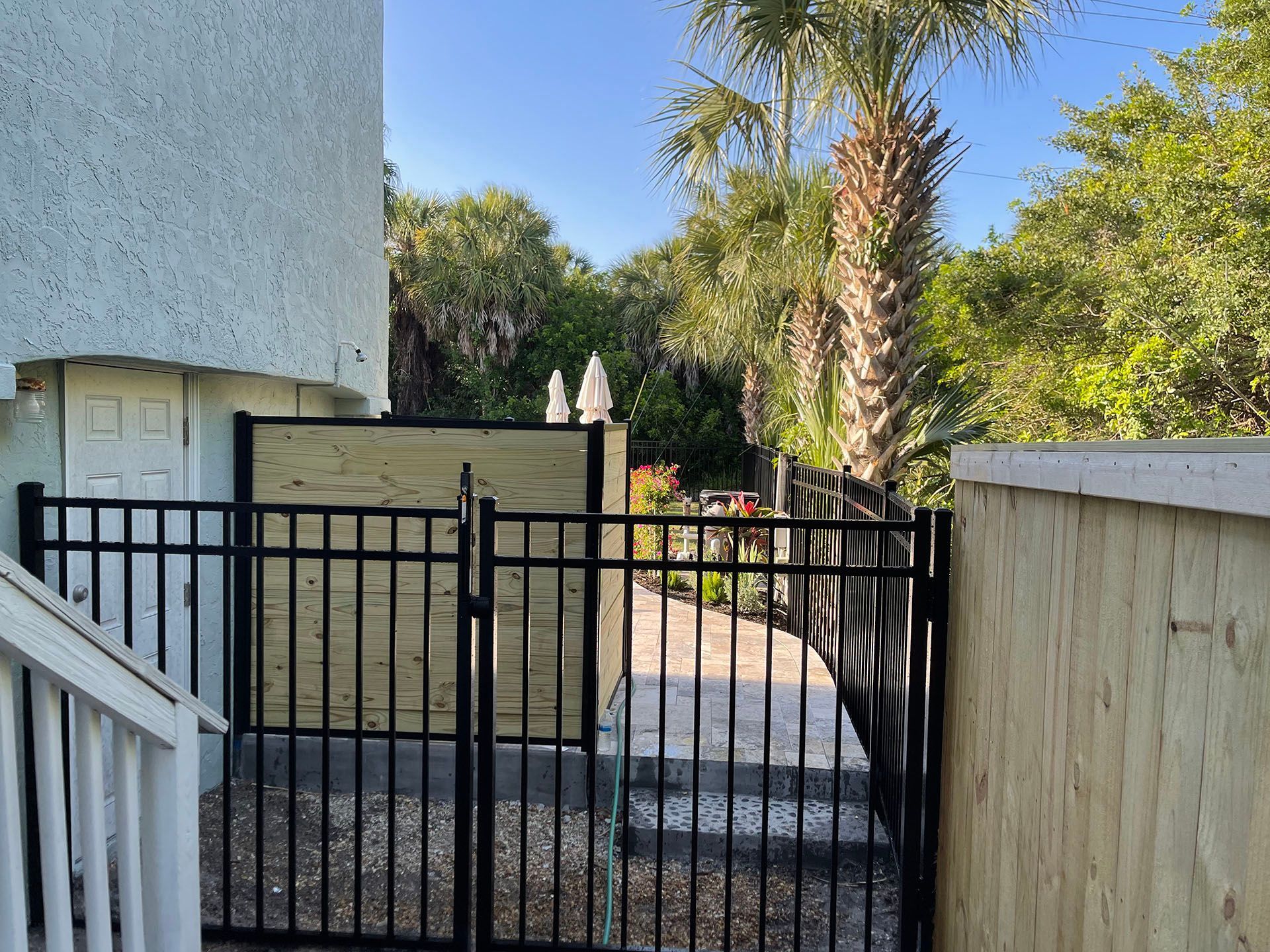 Black metal gate and fence lead to a pathway with trees; building on the left.