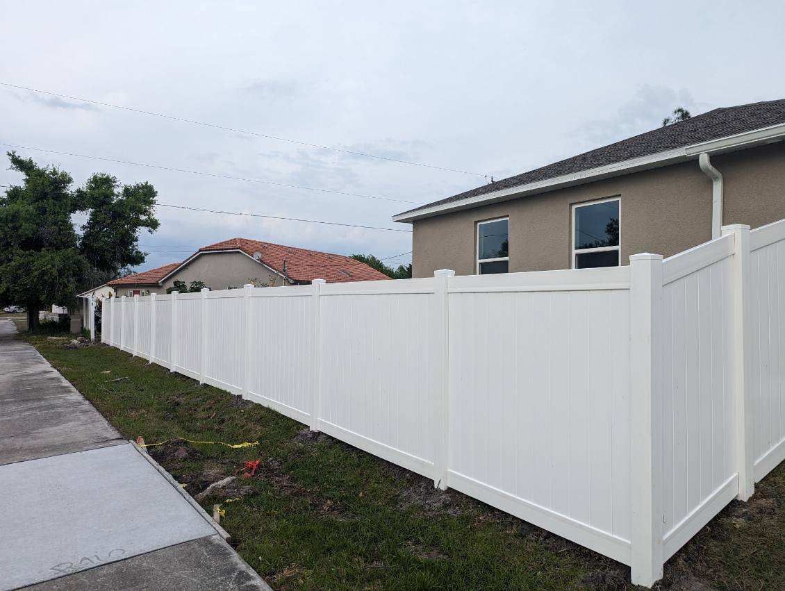 White vinyl fence along a sidewalk and grass, adjacent to houses under an overcast sky.