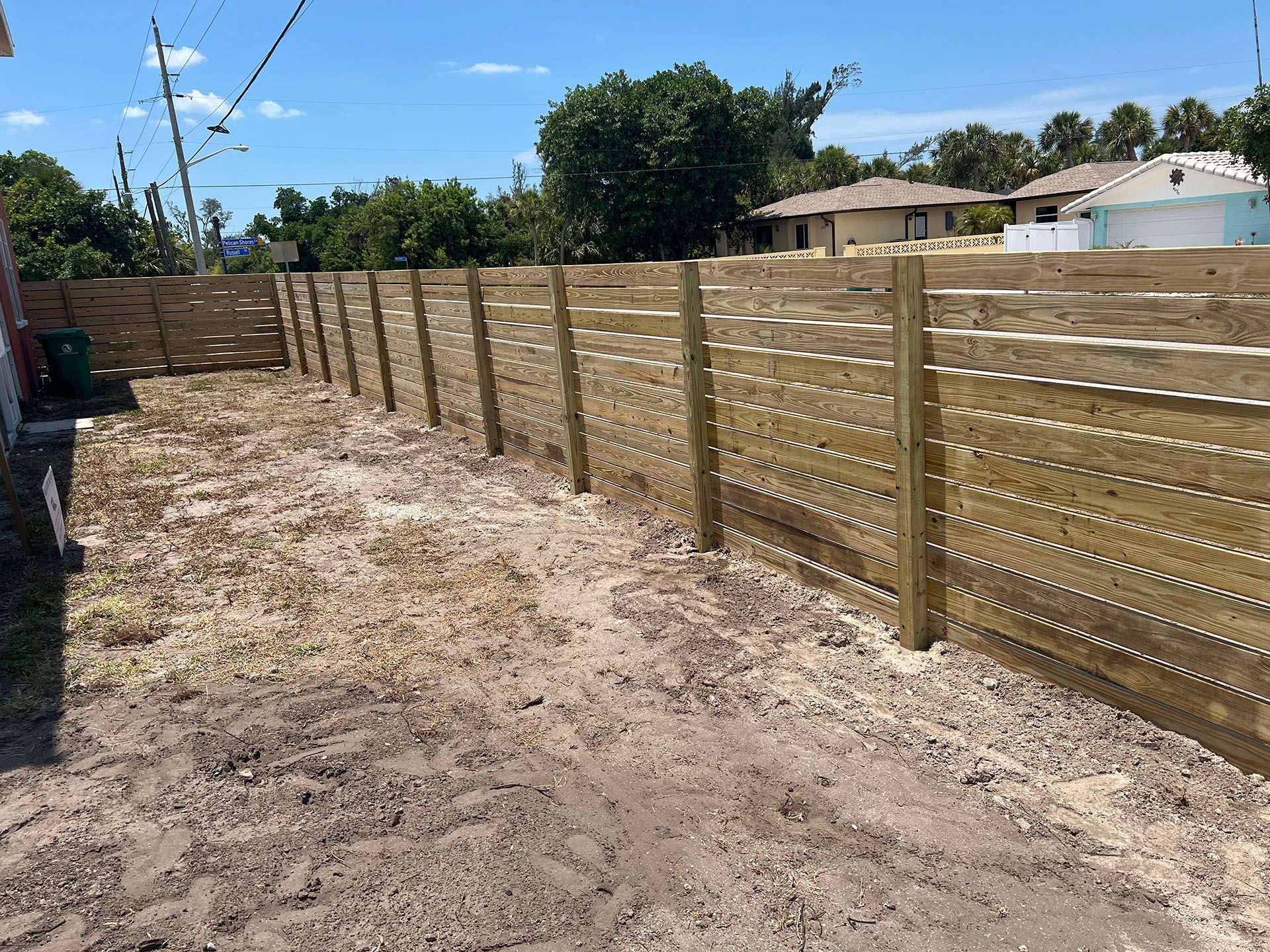 Wooden fence in a dirt yard, houses in the background on a sunny day.
