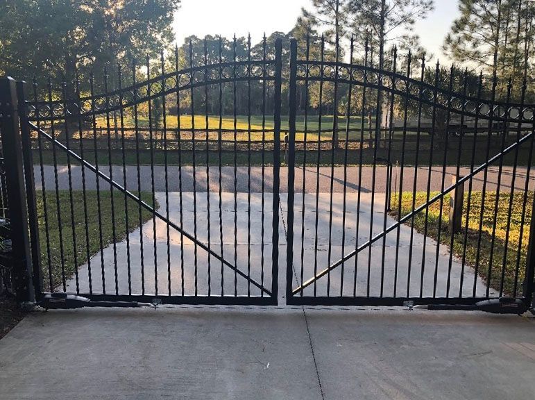 Black metal driveway gate on a concrete driveway, with a grassy area and trees visible in the background.