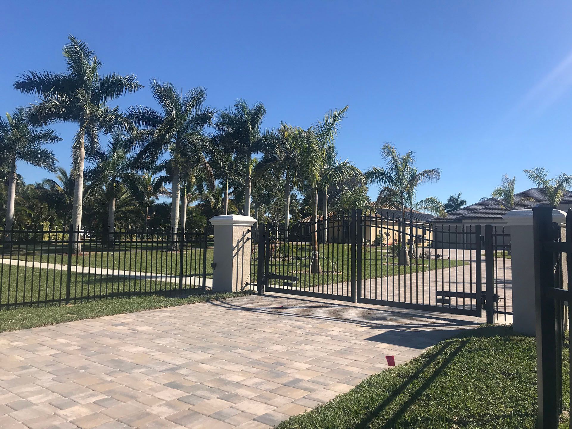 Black metal gate and fence on a brick driveway, leading to a yard with palm trees under a blue sky.