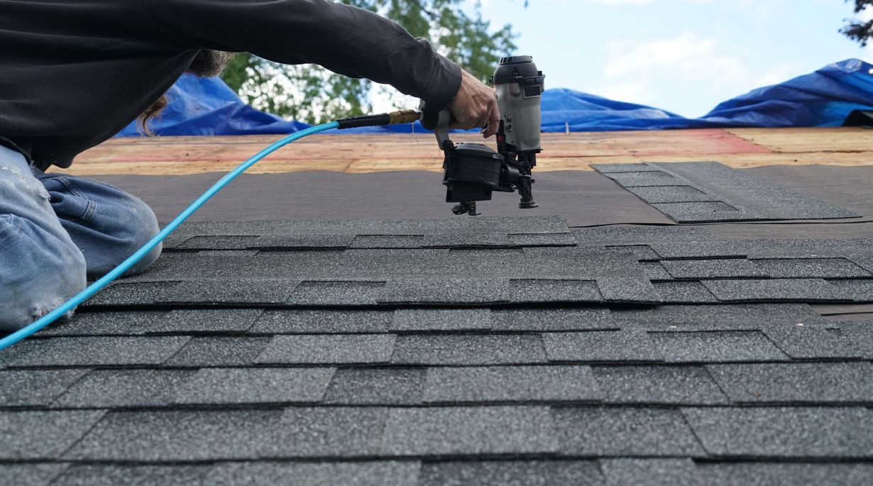 Roofer kneeling, using a nail gun to attach shingles on a gray asphalt shingle roof. Blue air hose visible.