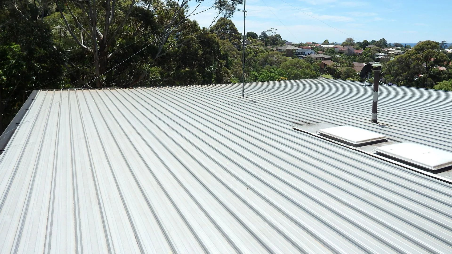 Metal roof with skylights, trees, and buildings in the background on a sunny day.