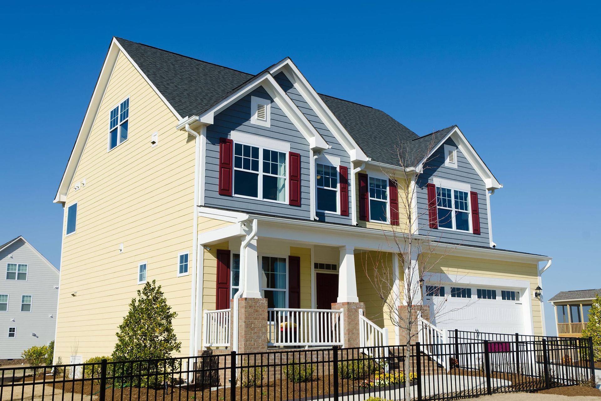 Yellow suburban house with white porch, red shutters, and black fence under a blue sky