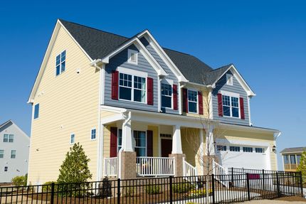 Two-story yellow house with white porch, red shutters, and a black fence under a blue sky