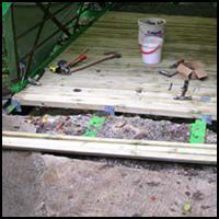 Wooden deck with tools and a paint bucket on top, above a dirt area and green fencing