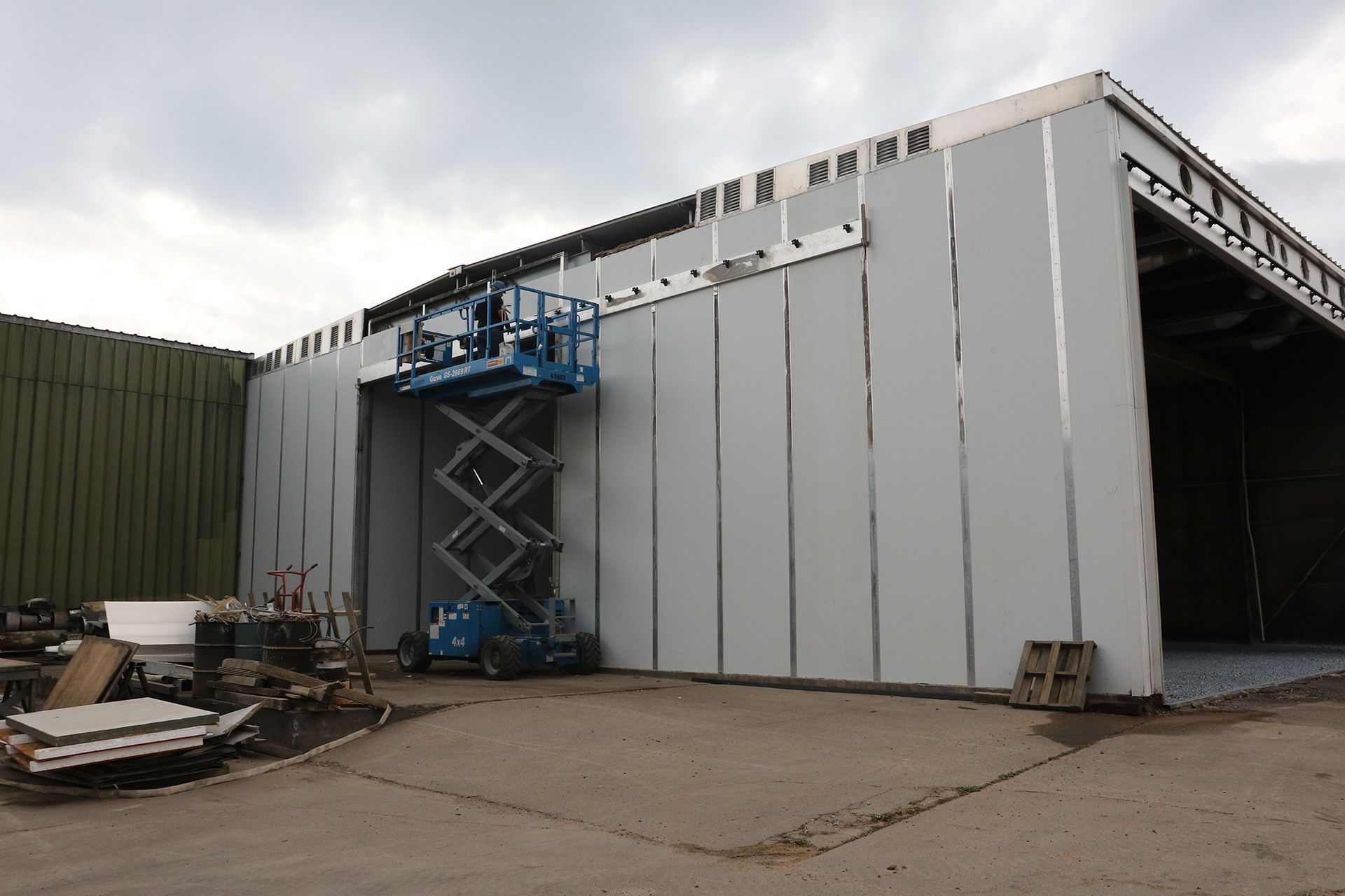 Large white industrial building with a blue scissor lift at the entrance and construction materials nearby