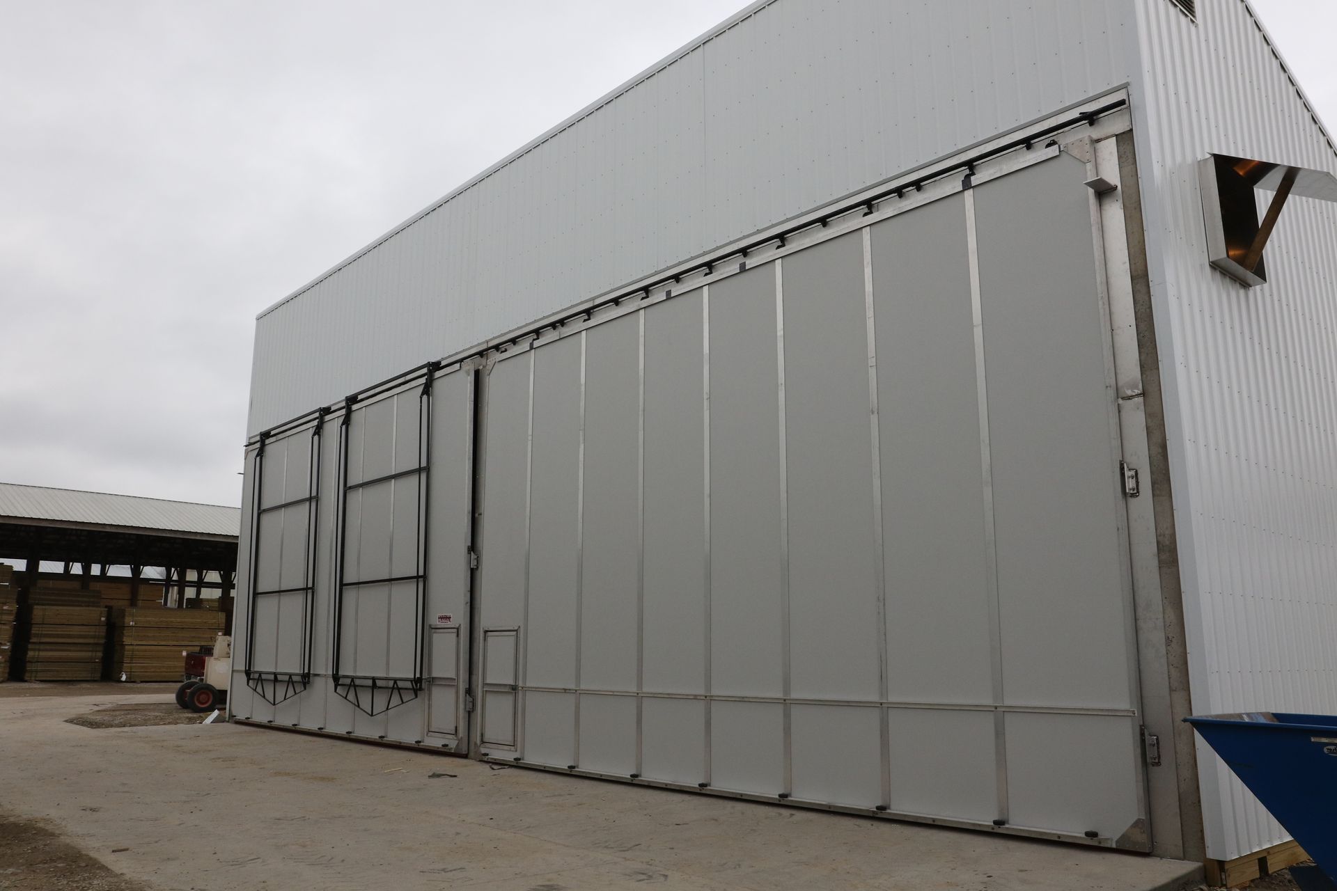 Large industrial metal building with a tall gray loading door on a gravel lot under an overcast sky