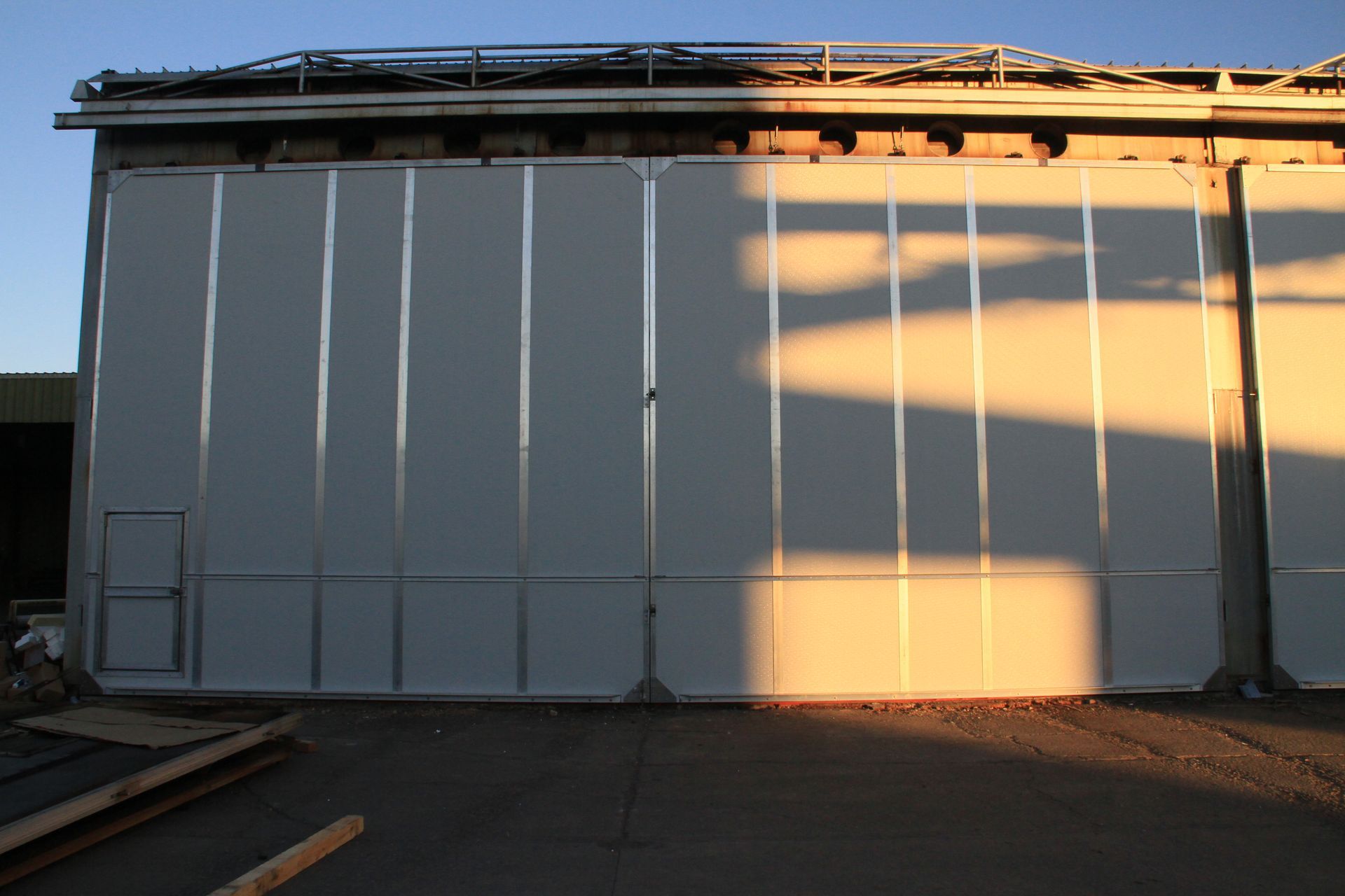White industrial building wall with roof, vertical seams, and sunlight casting shadows on the facade