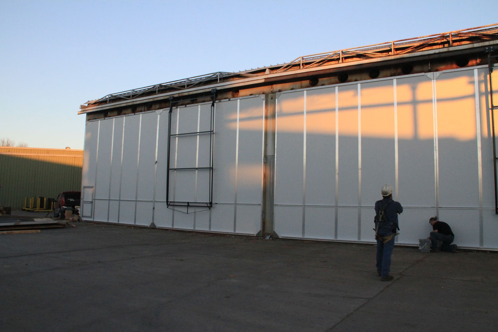 Large white industrial building at sunset with two people standing near the wall and roof equipment above
