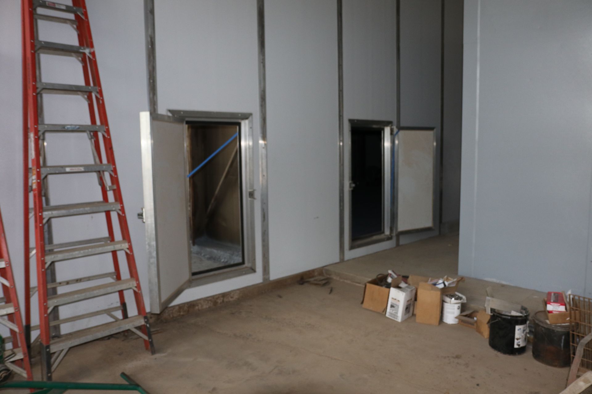 Interior construction site with gray walls, open wall cutouts, a red ladder, and buckets on the floor