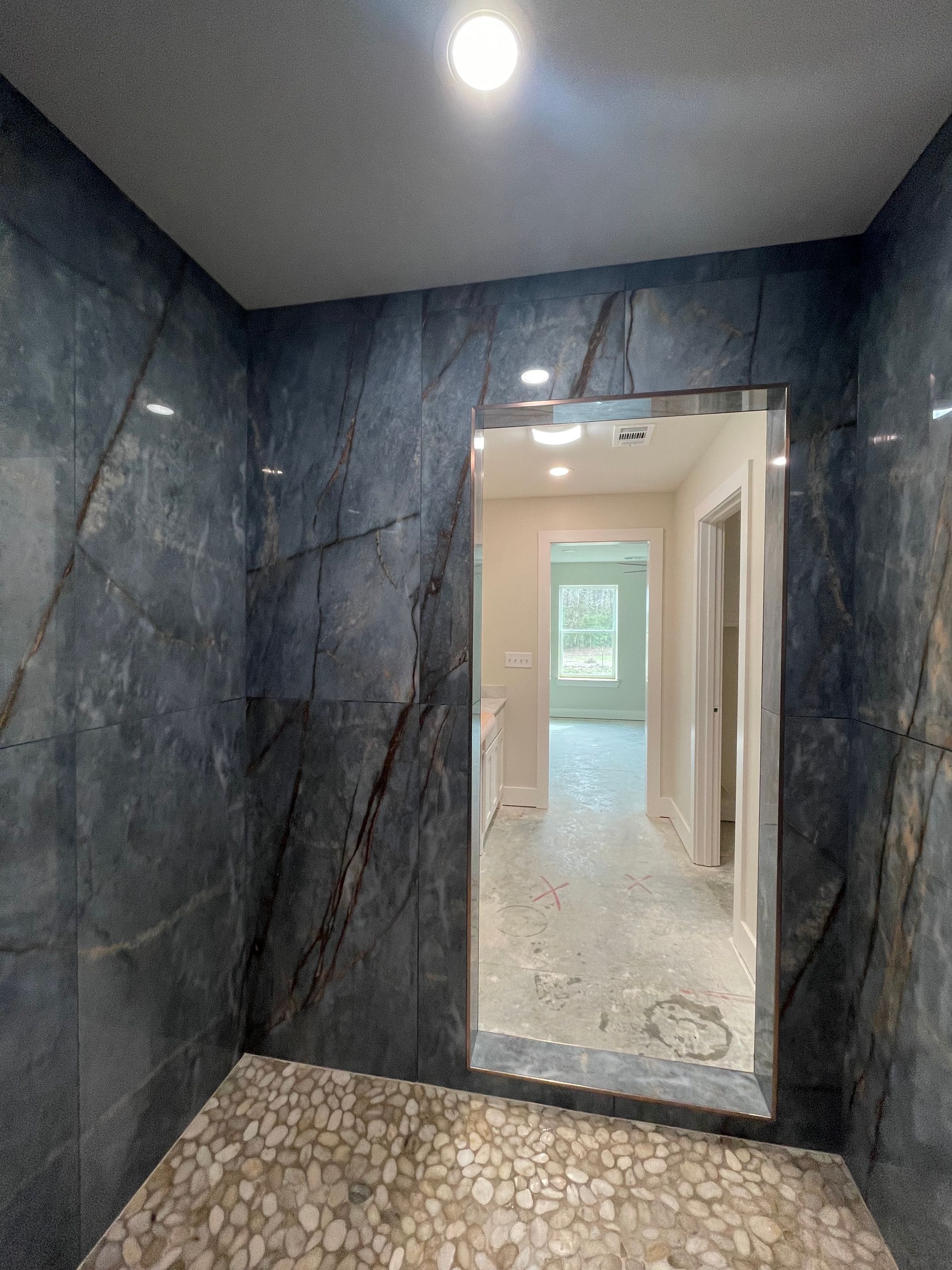 Dark tiled hallway with a wall mirror, recessed light, and a doorway to a bright room beyond.