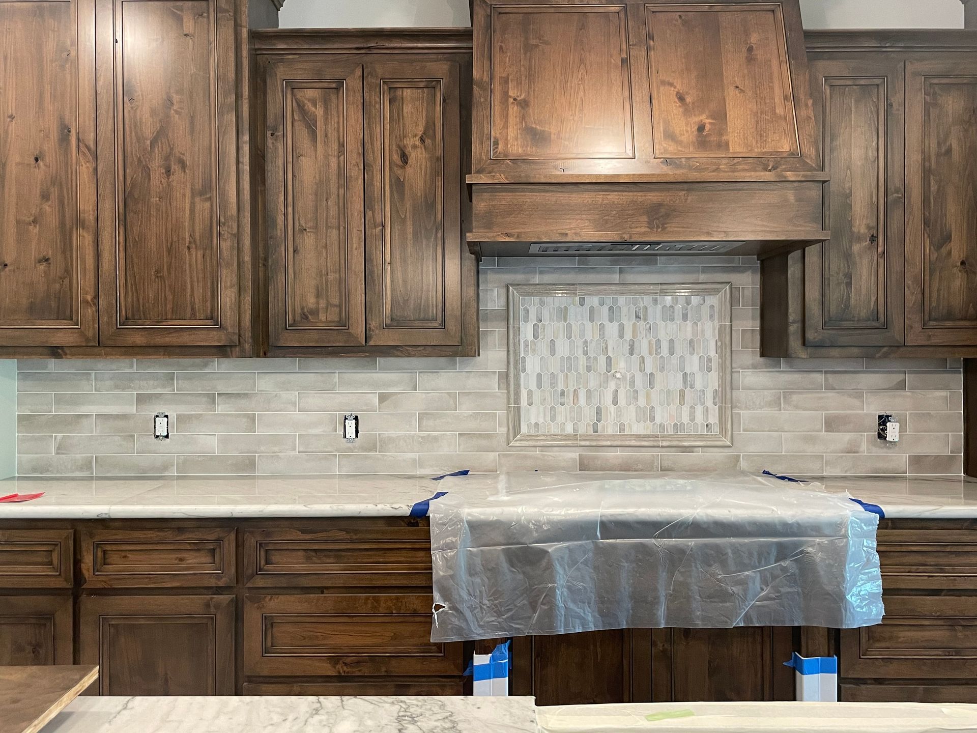 Kitchen with dark wood cabinets, beige tile backsplash, and a covered countertop under a range hood