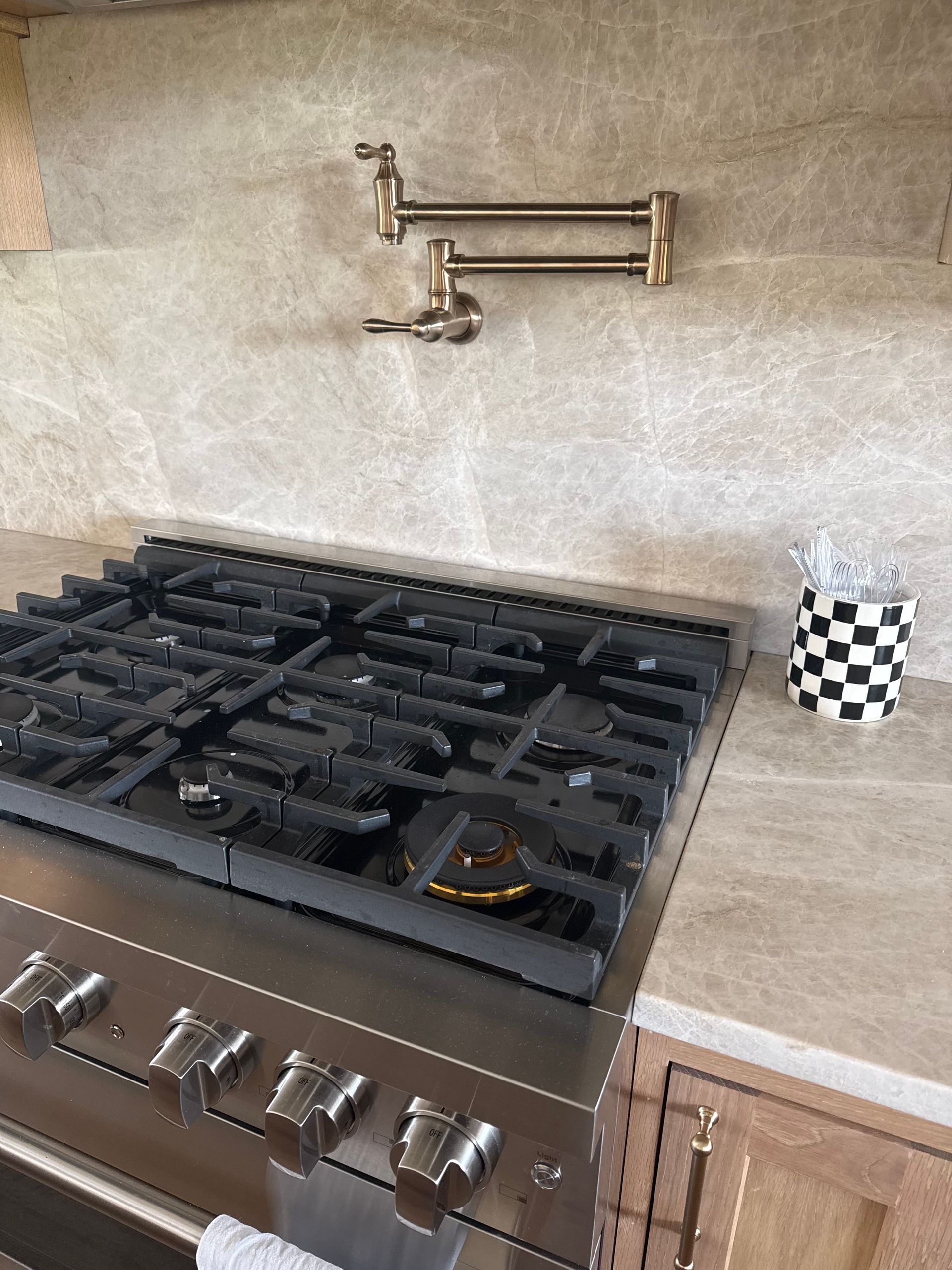 Stainless steel gas stovetop beneath a wall-mounted pot filler, with a checkered cup on the right counter.