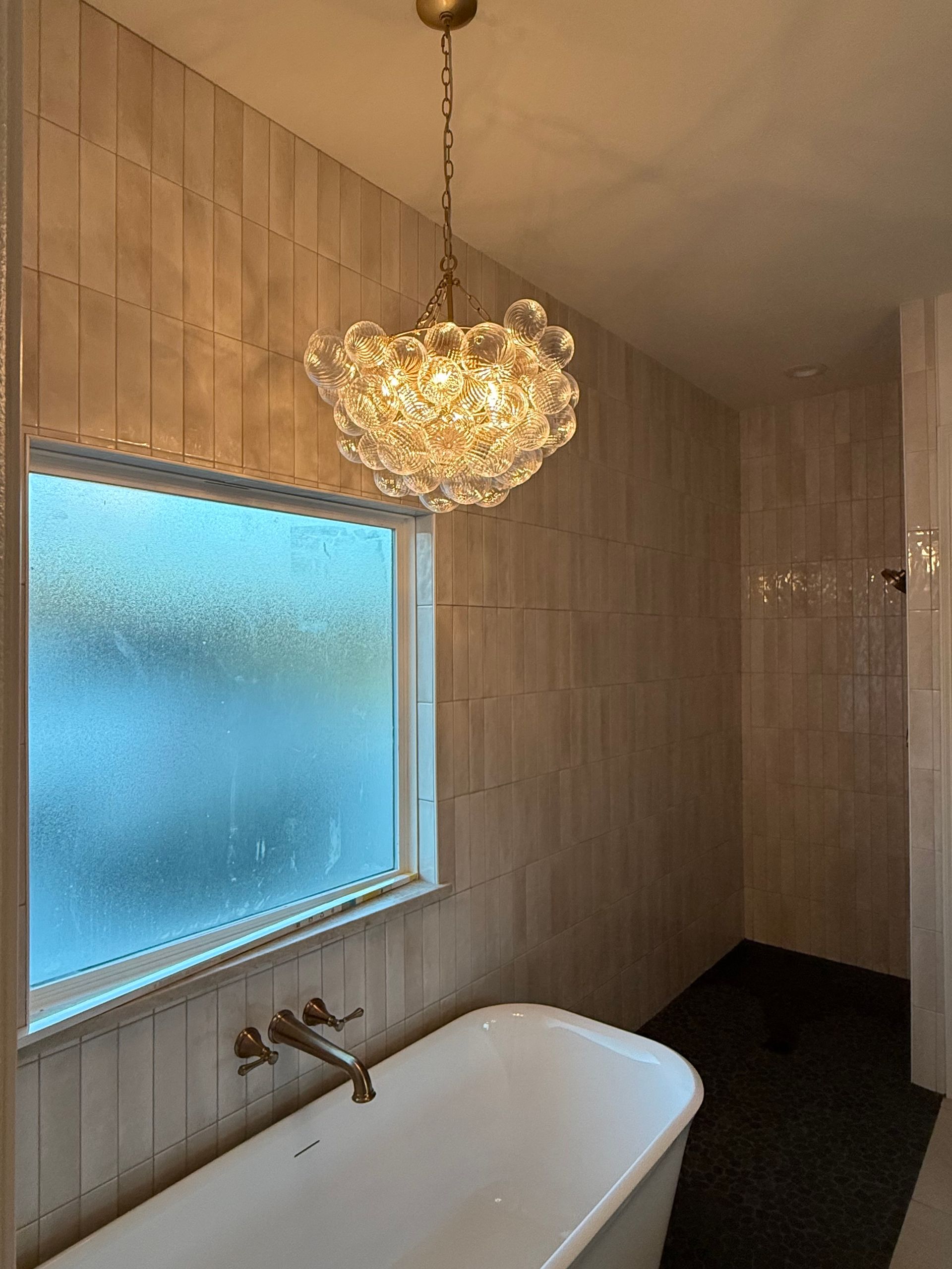 Modern bathroom with a freestanding tub, frosted window, wood-paneled walls, and a hanging chandelier.