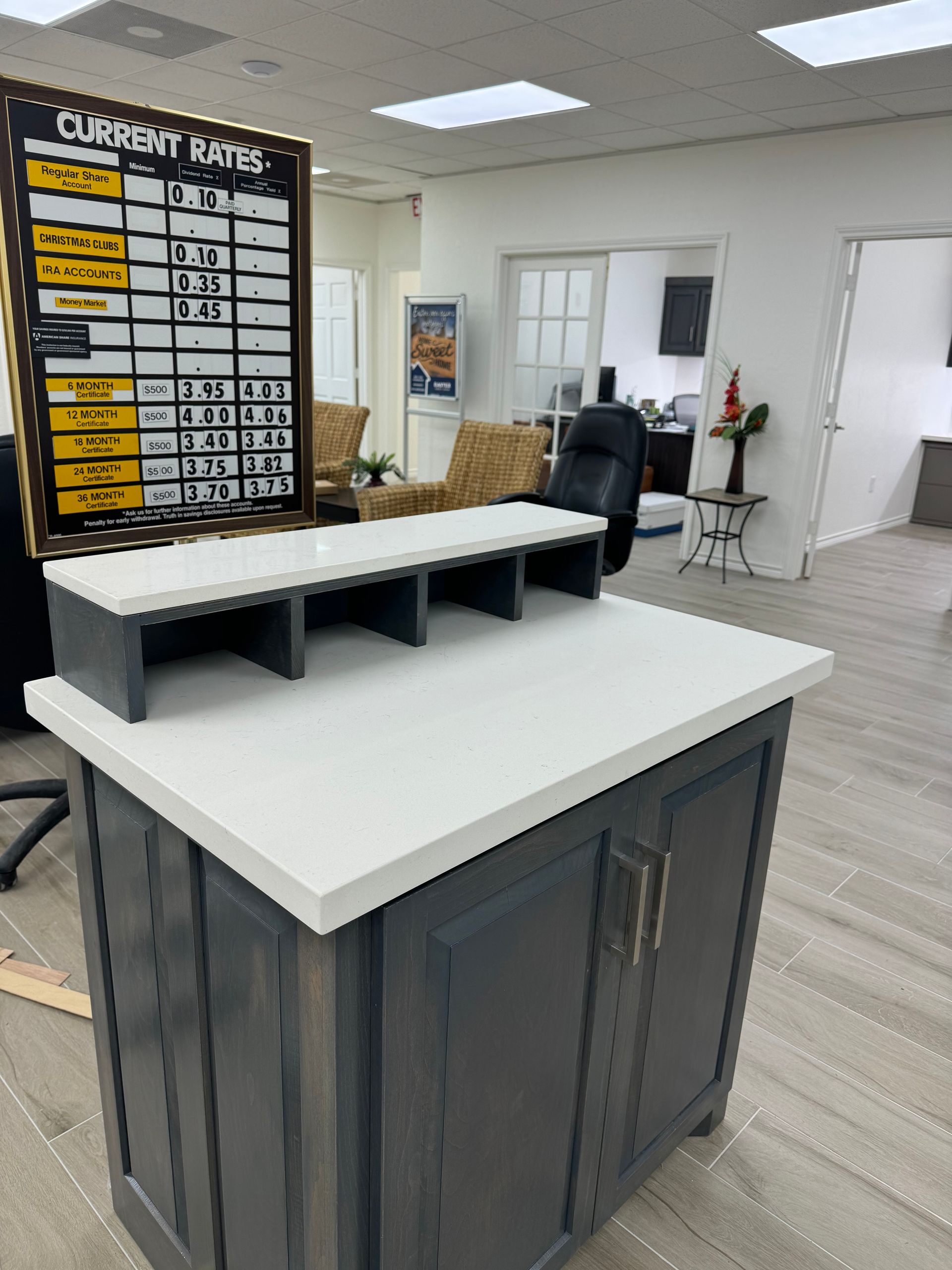 Office reception counter with white countertop, black cabinet, menu board, and potted plant in a bright room