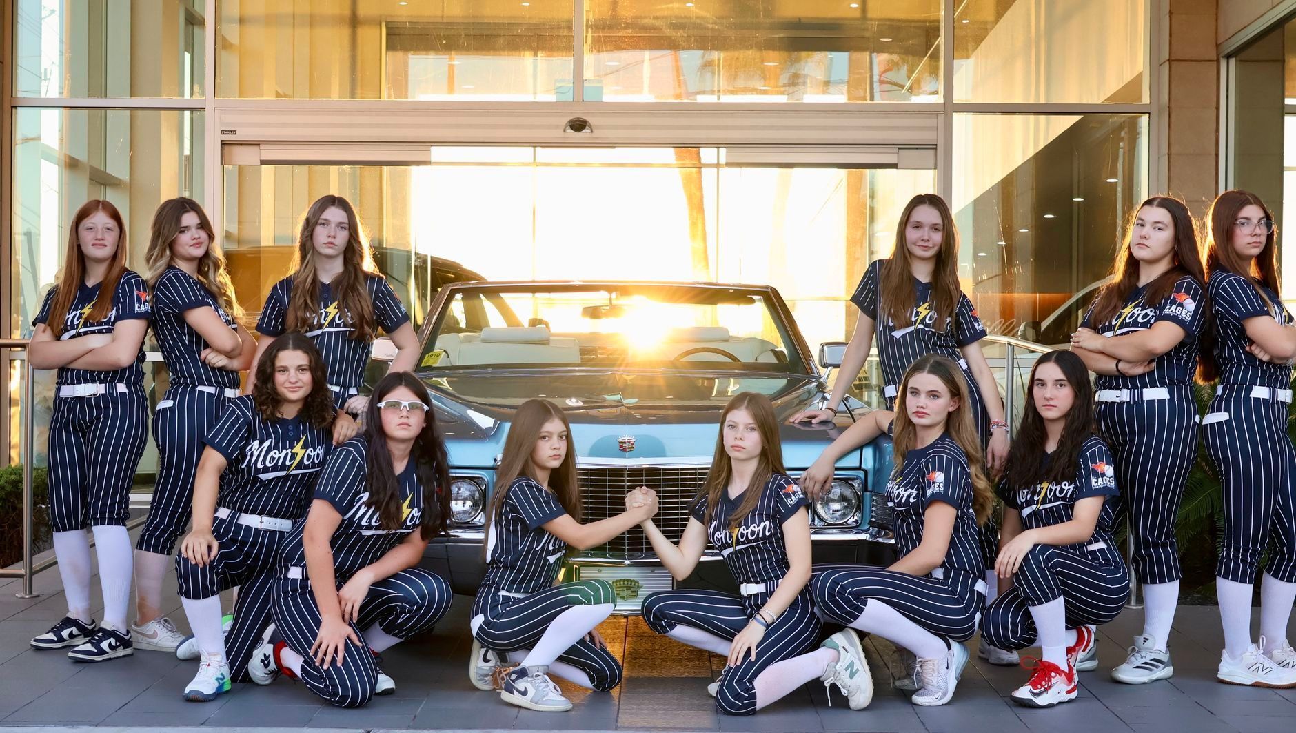 Softball team in navy and white uniforms poses by a light blue car; setting sun in background.