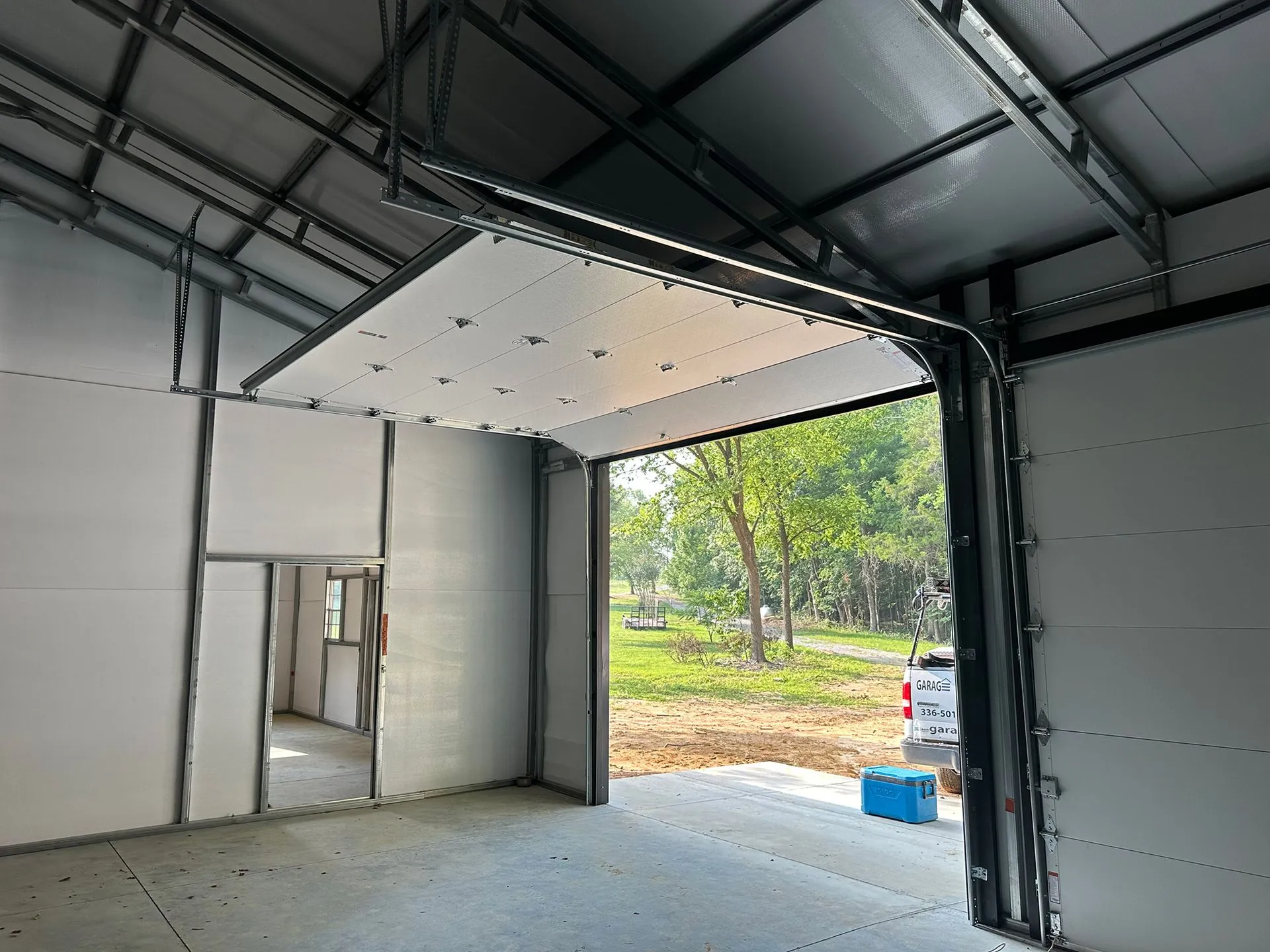 Interior view of a garage with an open door, showing a white panel garage door with a forest outside.