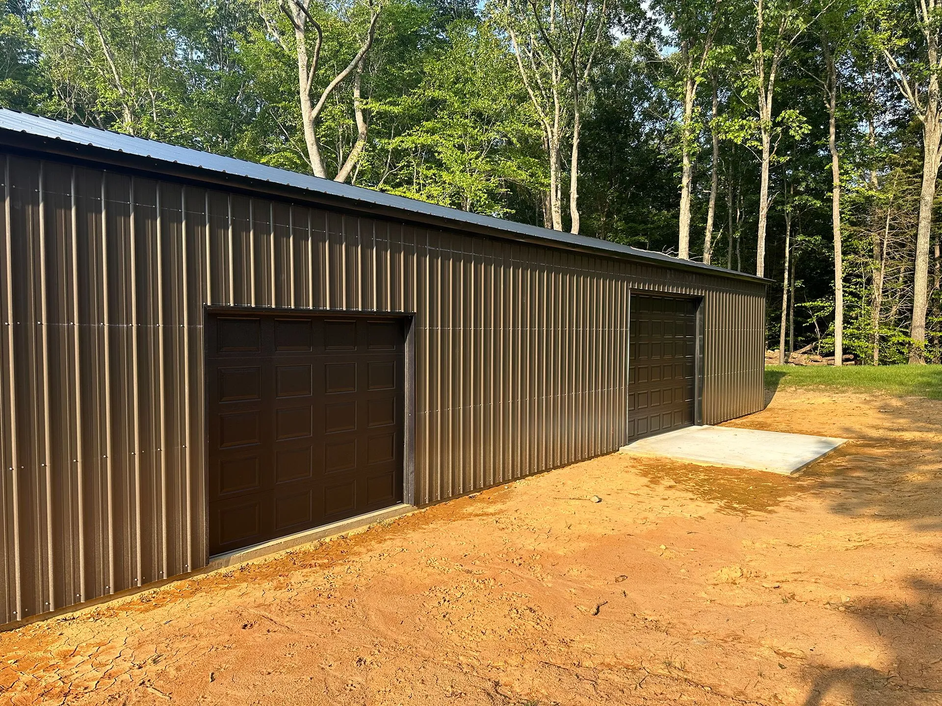 Brown metal shed with two dark doors, gravel ground, surrounded by trees.