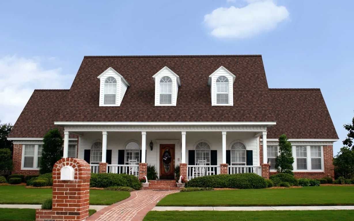 Two-story white house with brown roof and porch, brick walkway and mailbox, green lawn, clear sky.