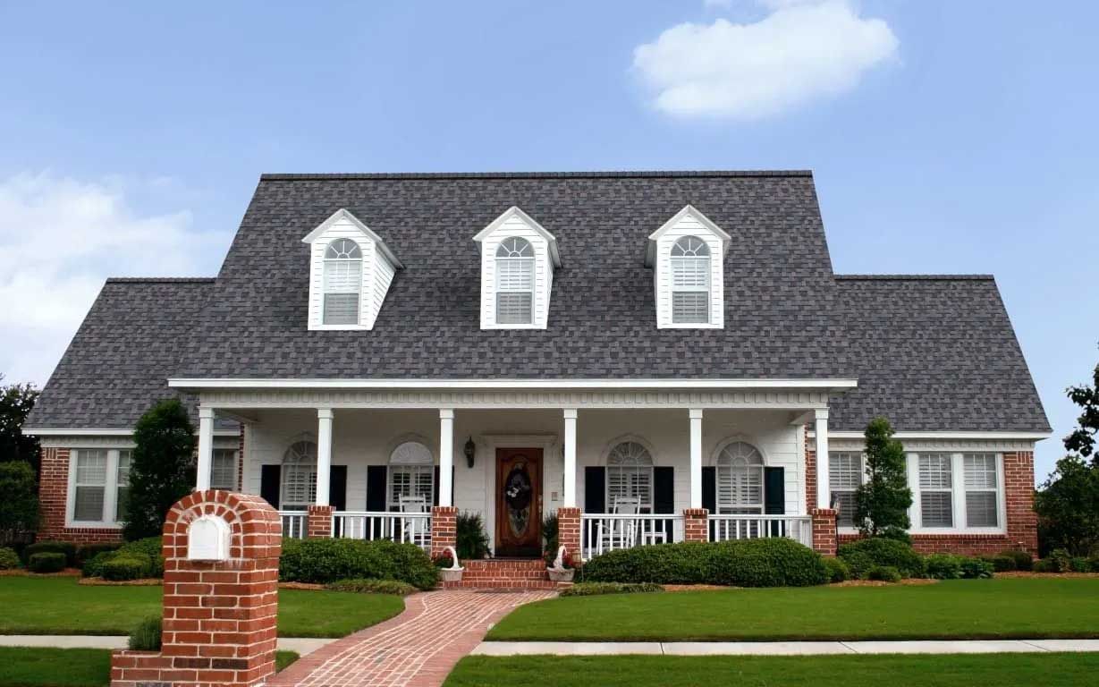 Brick house with porch, dormers, and brick walkway on a grassy lawn under a blue sky.