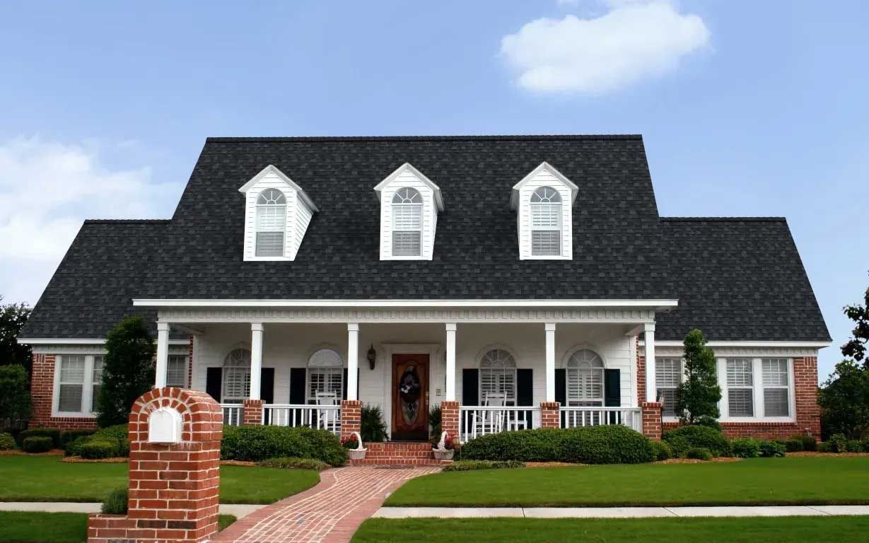 A two-story white house with a porch and dormer windows, red brick path and mailbox, green lawn, blue sky.