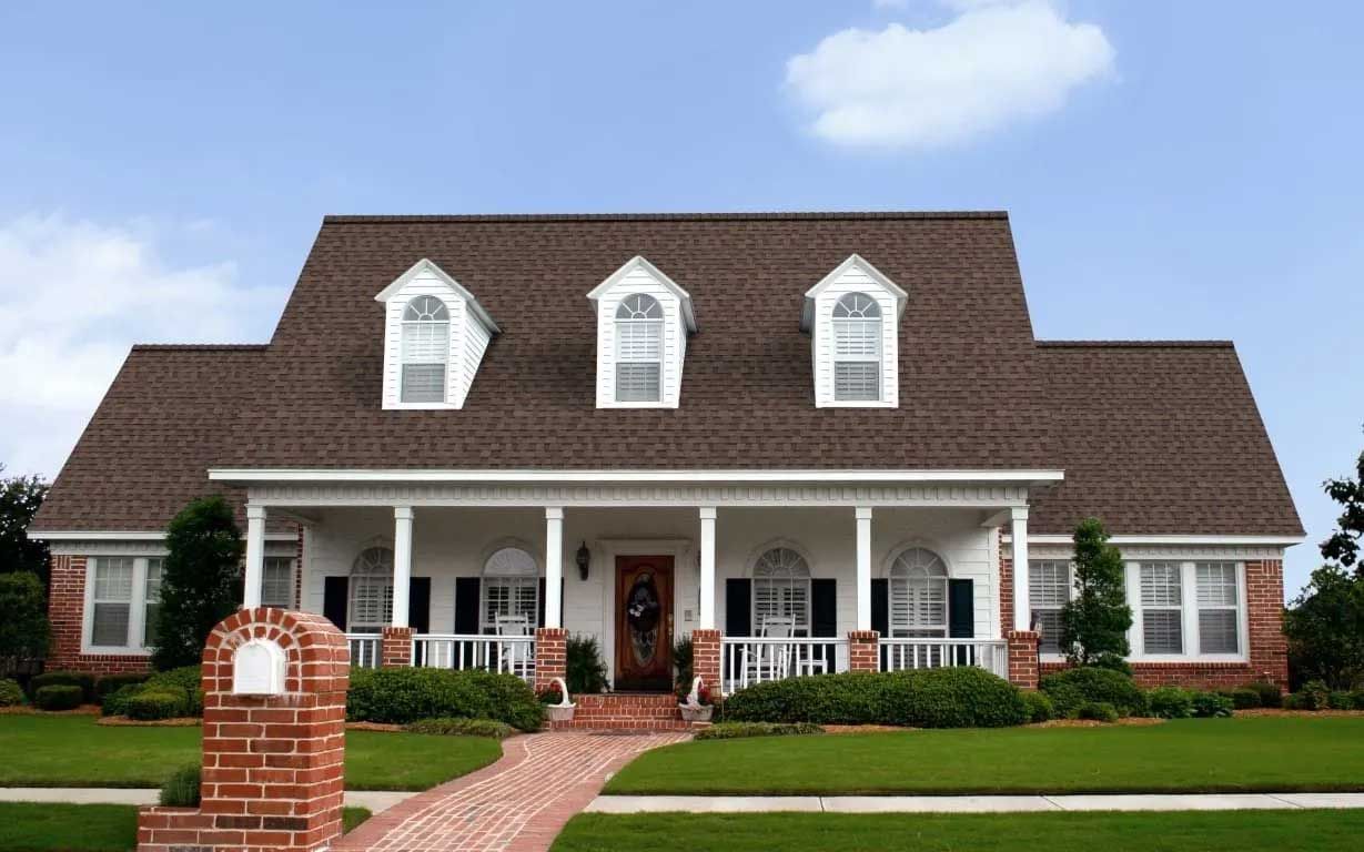 House with a brown roof, white porch, brick walkway, and green lawn under a blue sky.